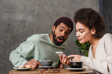 Amazed african american man sitting near girlfriend with smartphone and coffee in cafe