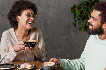 Cheerful african american couple with coffee talking near macaroons in cafe
