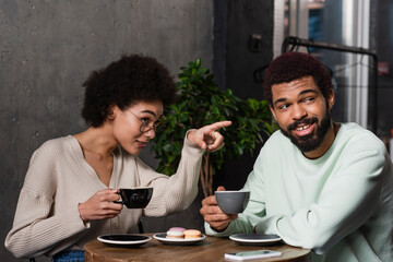 African american woman with coffee pointing with finger near smiling boyfriend in cafe