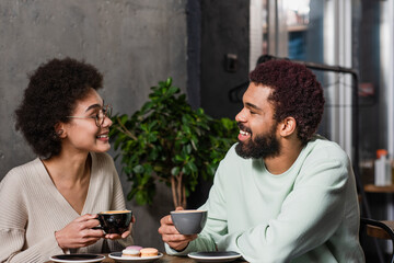 Side view of smiling african american couple holding coffee near macaroons in cafe