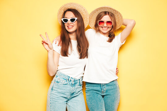 Two young beautiful smiling hipster female in trendy summer white t-shirt and jeans clothes.Sexy carefree women posing near yellow wall in studio.Positive and cheerful models in hats