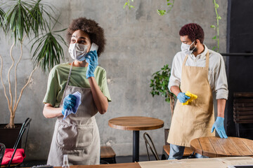 African american barista in medical mask and latex gloves holding rag and talking on smartphone in cafe
