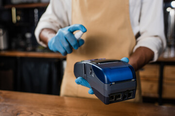 Cropped view of payment terminal and antiseptic in hands of african american barista in latex gloves