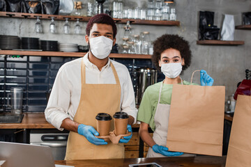 Fototapeta na wymiar African american baristas in medical masks holding paper bag and coffee to go in cafe