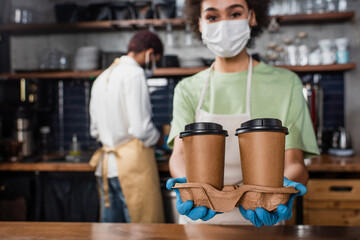 Paper cups in hands of african american barista in medical mask on blurred background