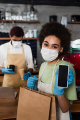 Fototapeta na wymiar African american barista in medical mask and latex gloves holding cellphone and paper bag in cafe