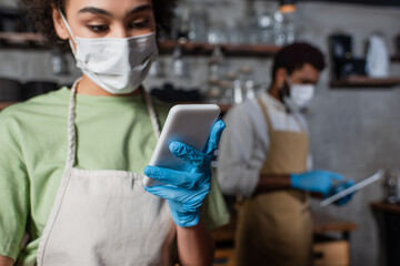 Fototapeta na wymiar Smartphone in hand of african american barista in medical mask on blurred background
