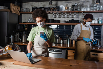 African american barista in medical mask using smartphone and laptop near colleague in cafe