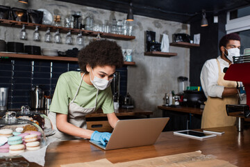 African american barista in medical mask using laptop near blurred colleague and coffee machine in cafe