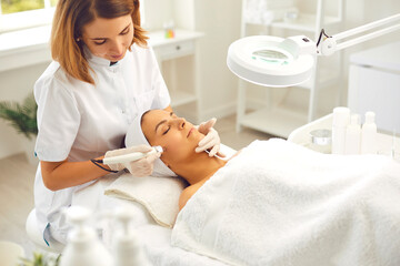 Obraz na płótnie Canvas Microdermabrasion skincare in beauty salon. Smiling woman dermatologist making skincare procedure of ultrasound Microdermabrasion with special metal machine for relaxing young woman