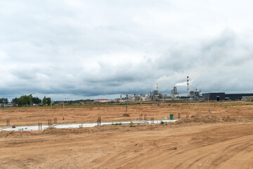 An empty construction site is located next to a large factory. Large factory chimneys emit thick, white, noxious smoke against the gray sky. Environmental disaster and the collapse of civilization.