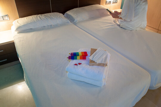 Woman sitting on the edge of a hotel bed consulting the mobile phone. With folded towels at the foot of the mask with a protective mask and detail of two red hearts and two gay flags