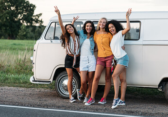 Happy diverse friends at camper van. Four smiling women standing together on the roadside near a car during vacation.
