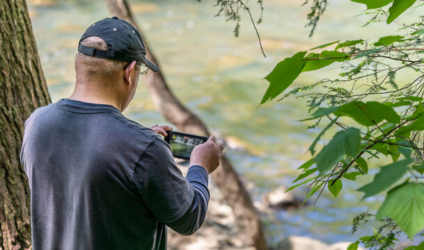 A Caucasian, middle age man takes a picture of a river with his cellphone. The man is wearing a blue shirt and hat