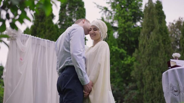 Medium shot of loving Caucasian man whispering on ear of smiling gorgeous Middle Eastern woman in wedding dress and hijab. Happy groom confessing love to bride at altar outdoors