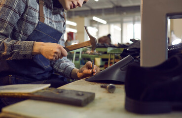 Shoe factory worker using professional tools to make new leather boots. Cropped shot woman sitting at workshop table working with hammer and nail. Manufacturing industry, footwear production concept