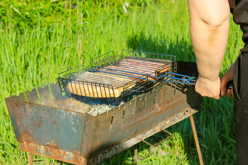 Raw sausage on a non-stick grill grid an the old metal BBQ. Preparing meat outdoors. Picnic in the woods on a sunny summer day. Green grass background. The process of cooking healthy food outdoors.