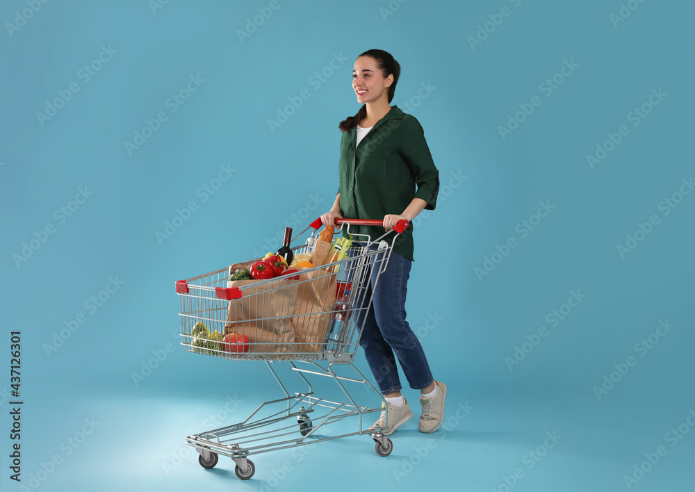 Poster Happy woman with shopping cart full of groceries on light blue background