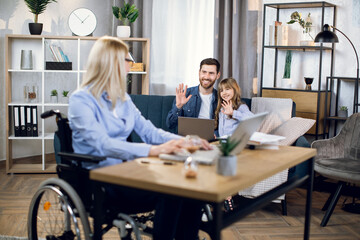 Young woman with disabilities using wireless laptop for remote work at home. Cute daughter and beloved husband sitting together on sofa and waving hands .