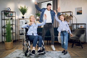 Happy woman in wheelchair dancing with beloved husband and pretty daughter at living room. Family spending actively time together. People with disabilities.