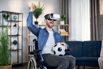 Joyful young man with disabilities playing virtual soccer match in 3D glasses. Handicapped bearded guy sitting in wheelchair and holding ball on knees.