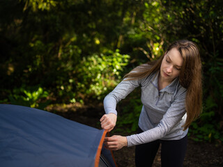 In the photo we see a bright sunny day. Dense green forest in the background. In the foreground, a beautiful young woman is laying out a tent. Woman resting in nature in the forest.