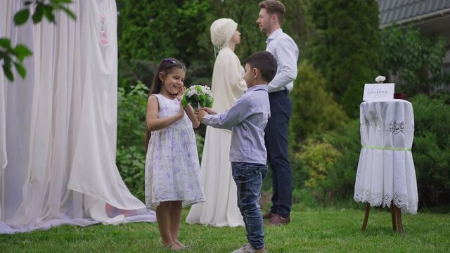 Cute Middle Eastern boy giving wedding bouquet to pretty girl on marriage ceremony outdoors. Cheerful children enjoying wedding celebration with blurred loving couple talking holding hands background