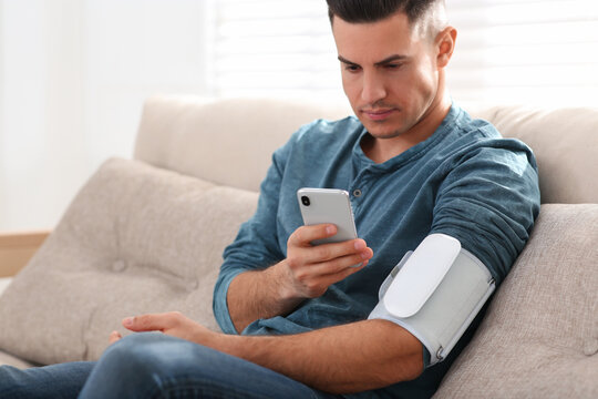 Man checking blood pressure with modern monitor and smartphone indoors