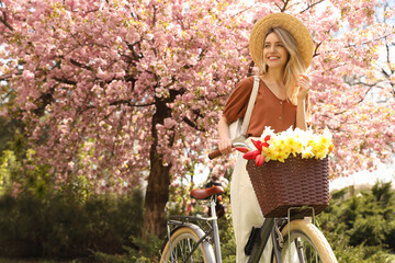 Beautiful young woman with bicycle and flowers in park on pleasant spring day. Space for text - obrazy, fototapety, plakaty