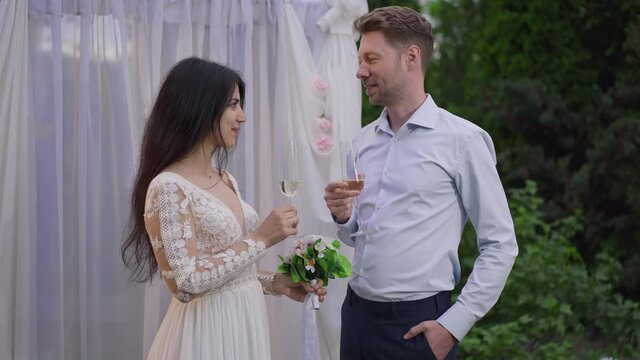 Happy interracial couple drinking champagne at wedding altar outdoors smiling. Portrait of loving positive Middle Eastern woman and Caucasian man getting married. Love and marriage concept