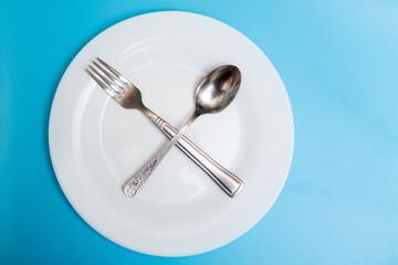 A spoon and a fork lie on an empty plate on a blue background. Diet concept, intermittent fasting.