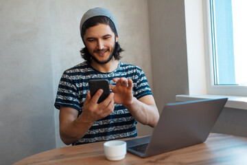 Portrait of young happy man using smartphone while working on laptop.