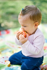 Little girl sits on a blanket and gnaws an apple. Close-up. Side view