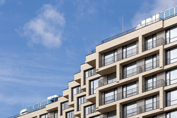 Fototapeta premium A fragment of a laconic residential high-rise building with balconies in light colors, with a blue sky in the background. Apartments in a modern skyscraper