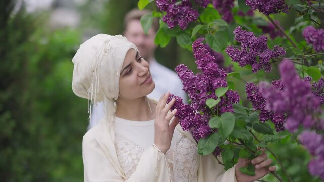 Medium shot of slim beautiful romantic Middle Eastern bride smelling blossom on tree branch with blurred unrecognizable groom at background. Happy woman and man in spring garden