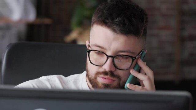 Close up Portrait. Handsome young guy worker in glasses speaking on the mobile phone and explaining something of working process while looking at the computer screen. Freelancer in home office.