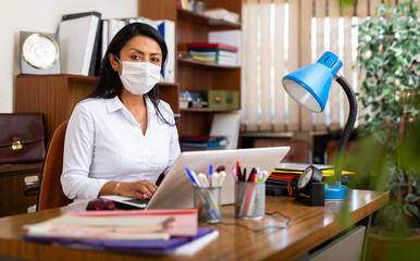 Fototapeta premium Woman in protective mask sitting at workplace with computer in her office