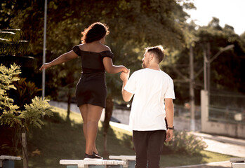 Boyfriend holding his girlfriend's hand to help her balance on a bench in a square.