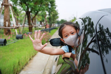 Asian little girl child wearing hygiene face mask pokes her head out of car window with show hand...