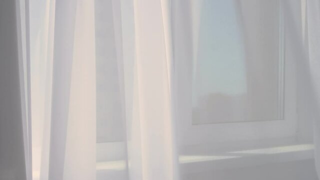 Hanging matte white tulle. The tulle is easily blown in different directions under the breeze. The curtain develops in the wind by the window.