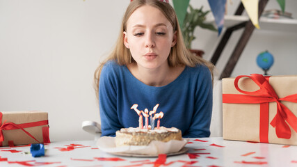 Obraz na płótnie Canvas Close-up of a Caucasian girl blows out the candles on a birthday cake. A young lady sitting at a table in the room is celebrating her birthday at home alone. Indoor party concept.