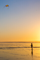 A lone figure flies a kite on the beach as the sun sets behind her turning the ocean and sky a beautiful bright orange.