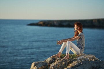 woman in white trousers and a T-shirt sits on a stone near the sea in the mountains