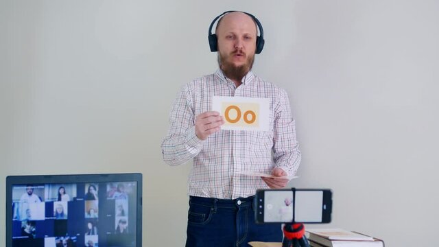 Learning English remotely online. A bald man with a beard gives a grammar lesson using flashcards to learn letters. Man in headphones and microphone