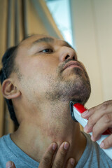 Asian man shaving beard with electric razor machine. Low angle view.