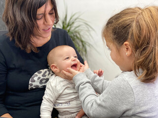 Little girl caressing her baby brother's face while he looks at her smiling. Mom holding baby.Caucasian family scene at home.