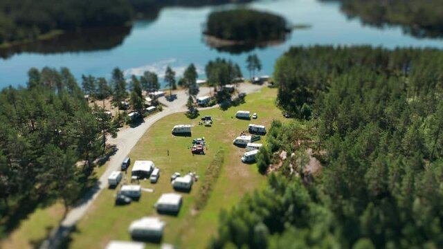 Tens of motorhomes, minivans, and cars parked in Kilefjorden camping are seen from the air on a sunny day. Camping is based on the Otra river and is surrounded by dense coniferous forest.