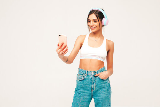 Beautiful smiling woman dressed in jeans clothes. Sexy carefree model listening music in wireless headphones. Adorable and positive female posing on grey background in studio.Taking selfie photos