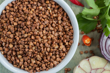 close up buckwheat with spices on a green wooden background. buckwheat in white plate with garlic, onion, parsley, and chili. Flat lay.