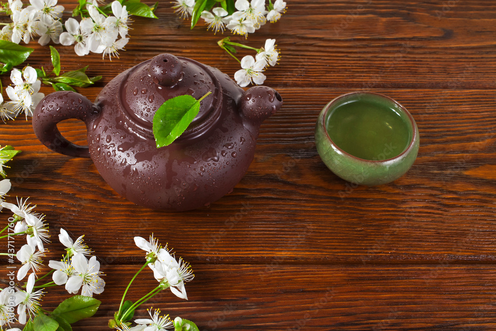 Wall mural Clay teapot and green cup of green tea with cherry flowers on a wooden background. 